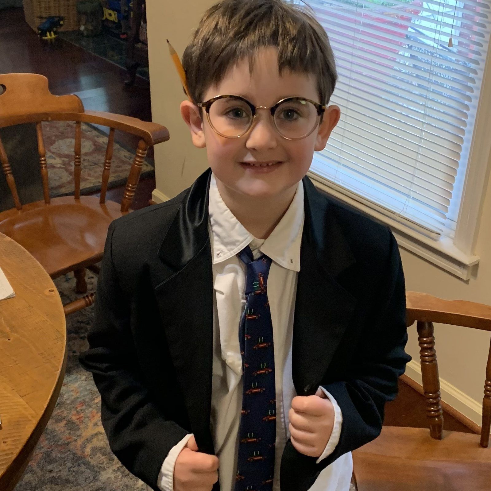 A young boy in glasses and an oversized blazer, grinning with a pencil tucked behind his ear.
